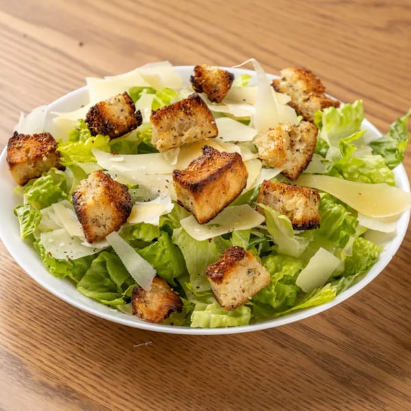 A Caesar salad with crisp green lettuce, shaved Parmesan cheese, and golden-brown croutons in a white bowl.
