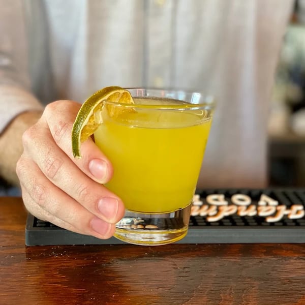 A hand holding a yellow cocktail garnished with a lime slice over a wooden bar.